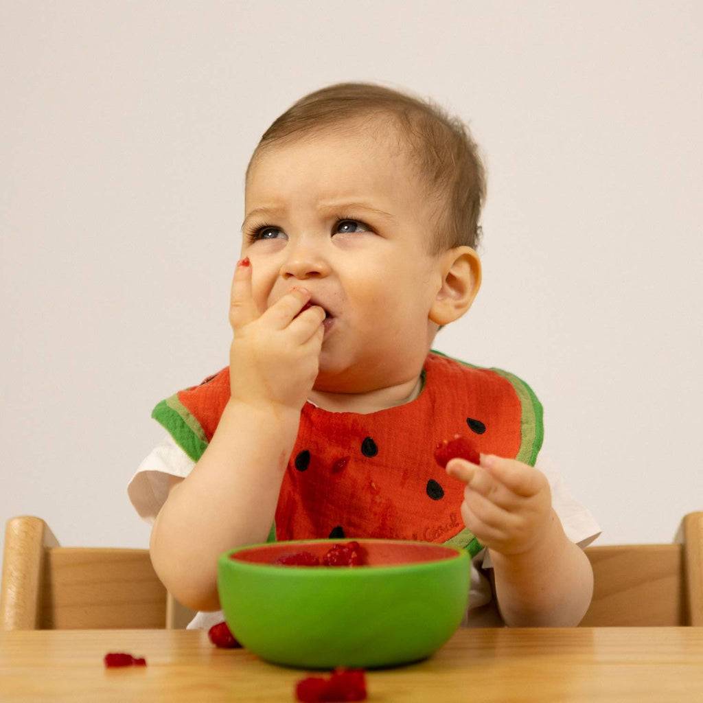 A baby with fruit on their hand sits at a table, eating from the non-toxic Wally the Watermelon Bowl & Spoon Set, wearing a curious expression.