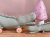 The Pull-Along Croc, a green knitted crocodile toy on wooden wheels from Egmont Toys, sits on a wooden surface next to another flat crocodile toy and a pink mushroom-shaped lamp with small holes.