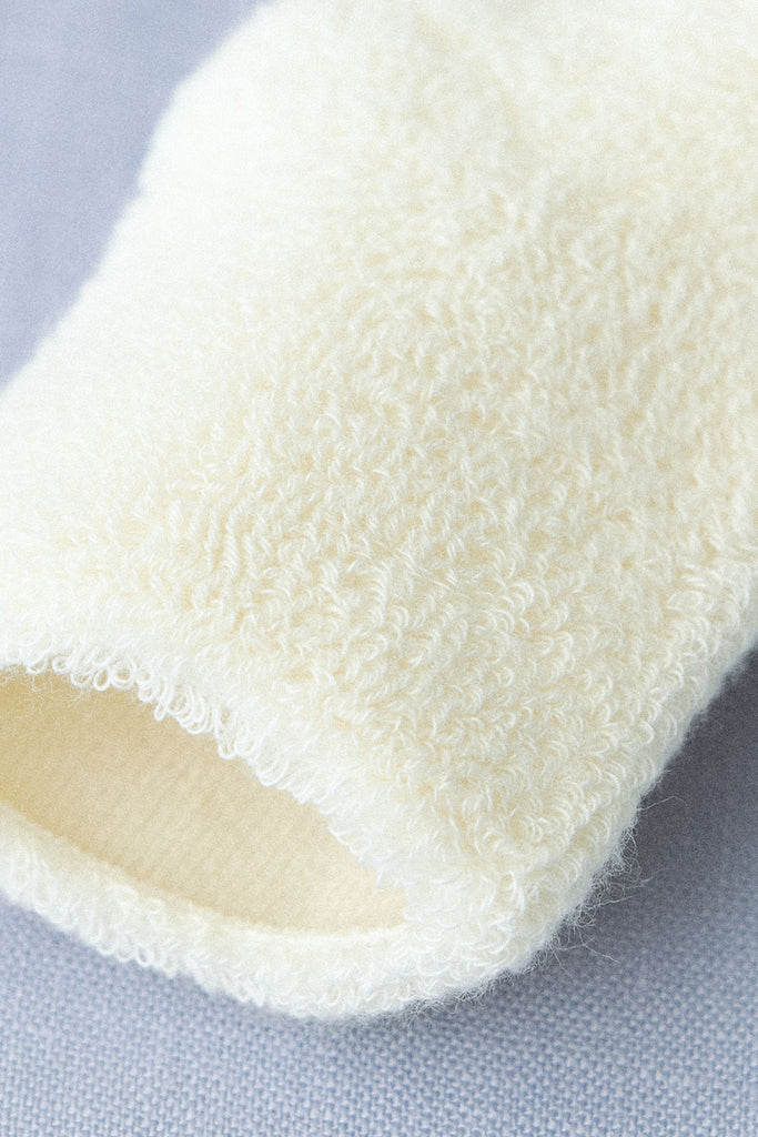 Close-up of a white, textured fabric with a fuzzy, looped surface, resembling terry cloth. The fabric appears to be soft and cozy, possibly part of Baby Pile Knit Socks - Natural or other Lithuania baby accessories, with the texture creating a visually interesting pattern. The background is a plain light blue surface.