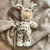 An Olli Ella Cozy Dinkum - Dog Domino plush toy, with closed eyes, a small smile, and an embroidered rainbow on its chest, lies on a beige textured blanket dressed in a spotted dog costume with floppy ears.