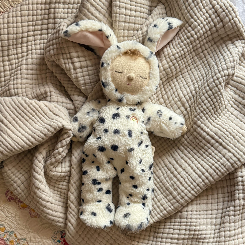 An Olli Ella Cozy Dinkum - Dog Domino plush toy, with closed eyes, a small smile, and an embroidered rainbow on its chest, lies on a beige textured blanket dressed in a spotted dog costume with floppy ears.