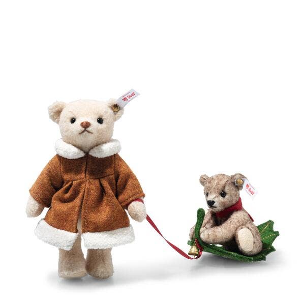 The Steiff Mama Teddy Bear With Child features a mama bear in a brown coat with white trim holding a red leash, leading her child bear on a green sled—ideal for your Christmas display against a white background.