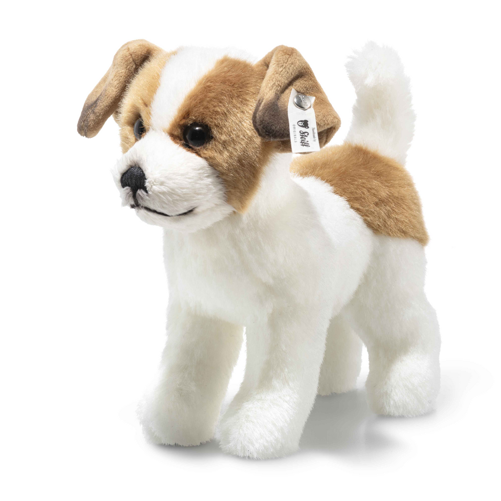 A Steiff Terrie Dog plush with white and brown fur stands on all four legs. This realistic stuffed dog features floppy ears, black eyes, a black nose, and a fabric tag attached to its left ear. The background is plain white.