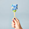 A hand with blue-painted nails holds the Garden Seed Pop (Single) by Modern Sprout, a lollipop-shaped gardening gift with a wooden stick and colorful wrapper—perfect for starting your own vegetable garden.