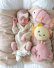 A newborn sleeps on a striped pillow, wearing a white outfit and bow headband. Next to the baby is the Cuddle + Kind Baby Fish, along with heirloom-quality sun and crab stuffed animals.