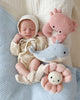 A newborn baby in a knit bonnet and cream outfit lies on a blue blanket, surrounded by heirloom-quality, hand-knit Cuddle + Kind Baby Pearl animals: a pink crab, blue whale, and pink smiling flower.