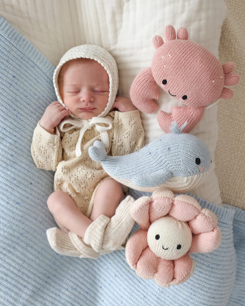 A newborn baby in a knit bonnet and cream outfit lies on a blue blanket, surrounded by heirloom-quality, hand-knit Cuddle + Kind Baby Pearl animals: a pink crab, blue whale, and pink smiling flower.