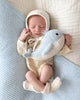 Newborn in a cream outfit and bonnet sleeps, cuddling an heirloom-quality Cuddle + Kind Baby Whale, on soft blue and cream blankets.