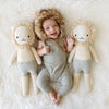 A joyful baby lying on a white blanket, wearing a lion hat and a gray onesie, flanked by two Cuddle + Kind Sawyer The Lion - Large dolls with serene expressions, crafted from Peruvian cotton