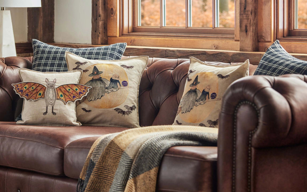 A brown leather sofa with a plaid blanket and Halloween-themed pillows, including the Coral & Tusk Bat Wing Surprise Pillow, sits before a sunlit window with wooden trim, topped off with a whimsical bat plush.
