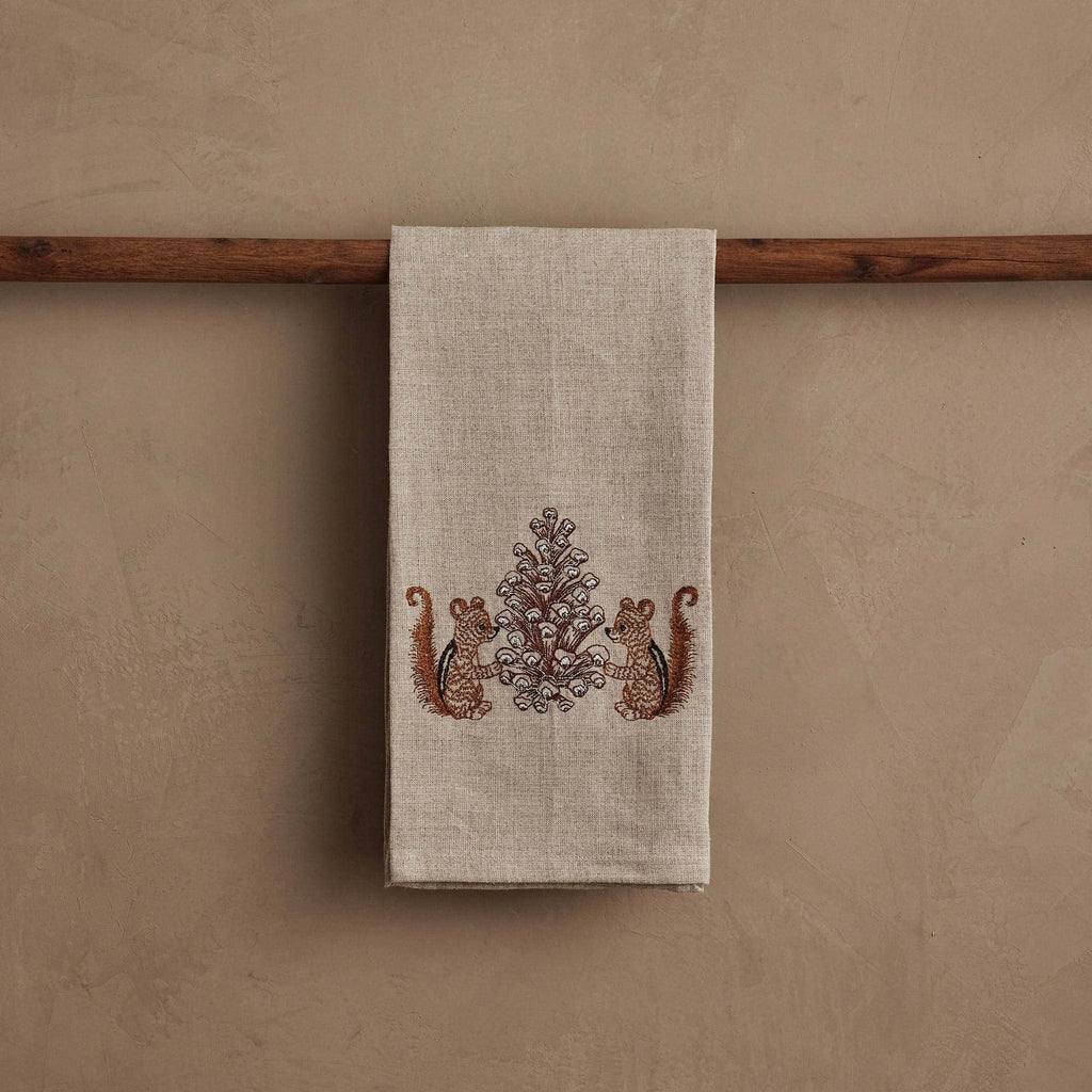 The Coral & Tusk Chipmunks with Pinecone Tea Towel in beige linen hangs on a wooden rod and features a woodsy design with two brown chipmunks facing a symmetrical, ornate pinecone tree pattern in the center.