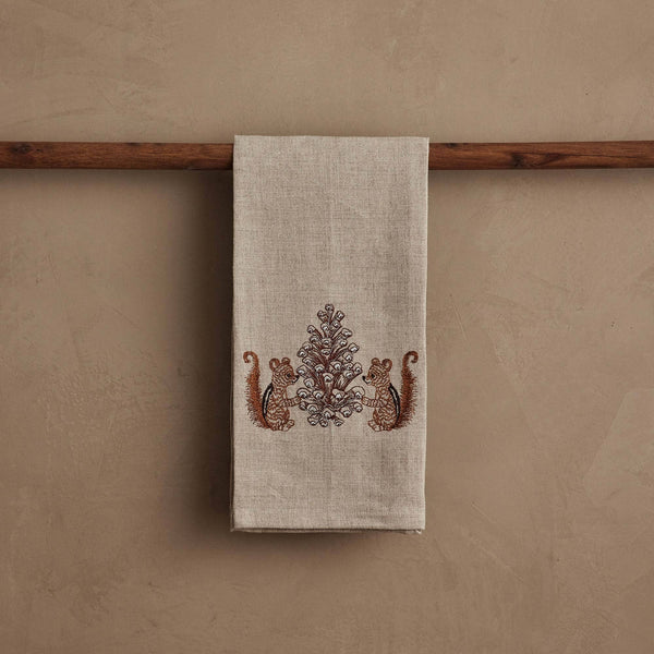 The Coral & Tusk Chipmunks with Pinecone Tea Towel in beige linen hangs on a wooden rod and features a woodsy design with two brown chipmunks facing a symmetrical, ornate pinecone tree pattern in the center.