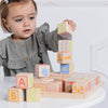 A young child in a gray dress carefully stacks colorful ABC Learning Blocks (ships in one week) on a white table, with more blocks scattered nearby.