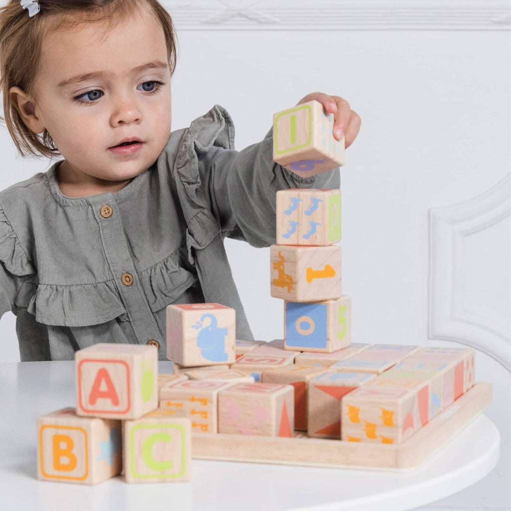 A young child in a gray dress carefully stacks colorful ABC Learning Blocks (ships in one week) on a white table, with more blocks scattered nearby.