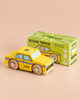 A yellow wooden toy taxi car, reminiscent of a classic New York taxi and labeled “TAXI,” sits in front of its green and yellow box, which reads “No. 39 GT245 Trixie | Made in England.” The car features checkered detailing and sturdy wooden wheels, making it the perfect toy for imaginative play.