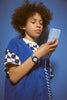 A child with curly hair, dressed in a blue vest and checkered shirt, wears the Mini Kyomo Watch - Blue Sunday - Blue Checkered and holds a retro corded phone against a blue background.