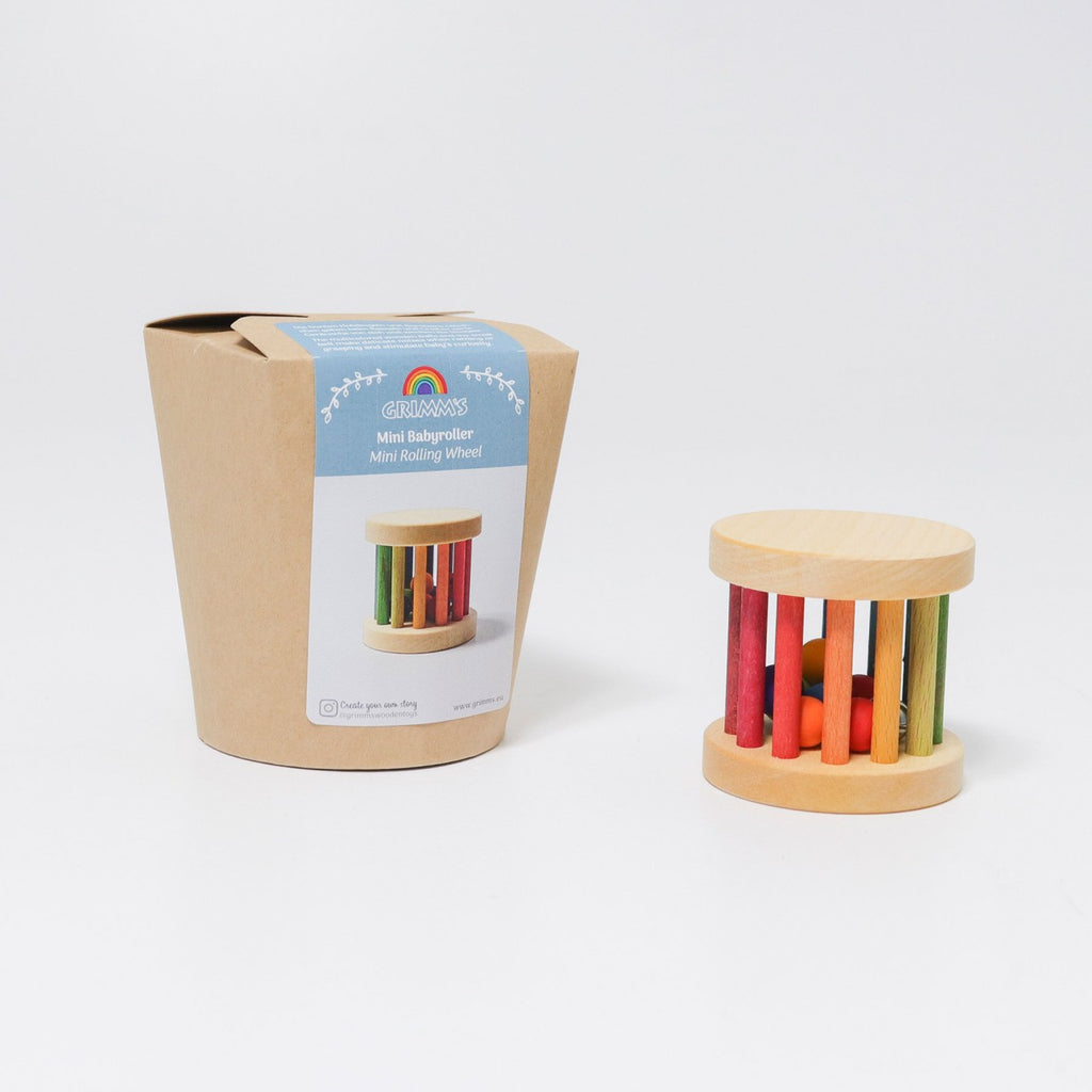 A small cylindrical wooden baby rattle toy with colorful bars and red balls inside, next to its cardboard packaging. The packaging has a colorful rainbow logo and text that reads, "Mini Rolling Wheel," featuring playful wooden beads in a vibrant design.