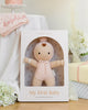 The Cuddle + Kind My First Baby (Blush), a soft hand-knit doll in a pink outfit, sits in its white box with flowers, a folded pink blanket, and a white cloth nearby. Ships in one week.