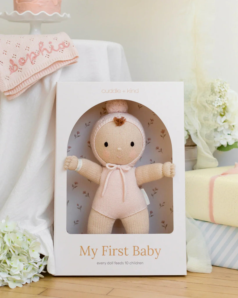 The Cuddle + Kind My First Baby (Blush), a soft hand-knit doll in a pink outfit, sits in its white box with flowers, a folded pink blanket, and a white cloth nearby. Ships in one week.