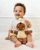 A smiling baby with curly hair sits on the floor, holding a Cuddle + Kind My First Baby (Buttercup) doll, made from natural cotton yarn with a yellow outfit and pom-pom hat. The baby wears a light shirt and striped shorts. Ships in one week.