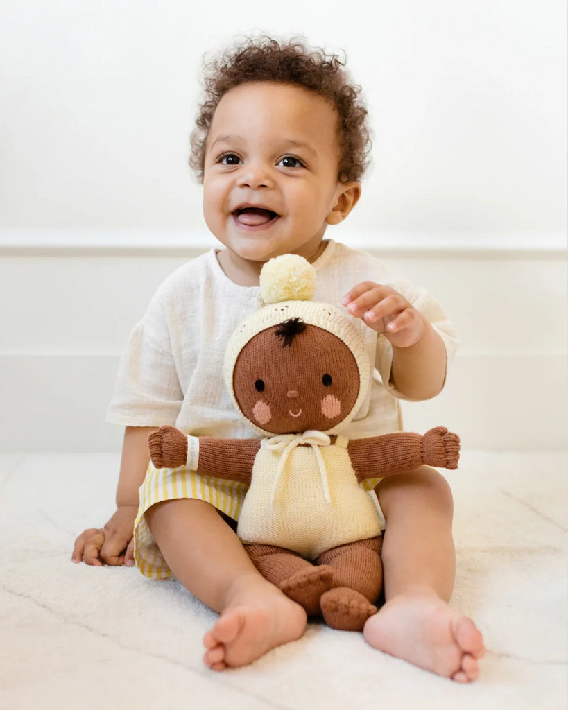A smiling baby with curly hair sits on the floor, holding a Cuddle + Kind My First Baby (Buttercup) doll, made from natural cotton yarn with a yellow outfit and pom-pom hat. The baby wears a light shirt and striped shorts. Ships in one week.