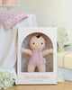 A boxed Cuddle + Kind My First Baby (Lavender) doll is displayed on a table. The doll wears a lavender outfit and bonnet. Near it are white flowers, a pink "Olivia" hat, and a pink cake on a stand. Ships in one week.