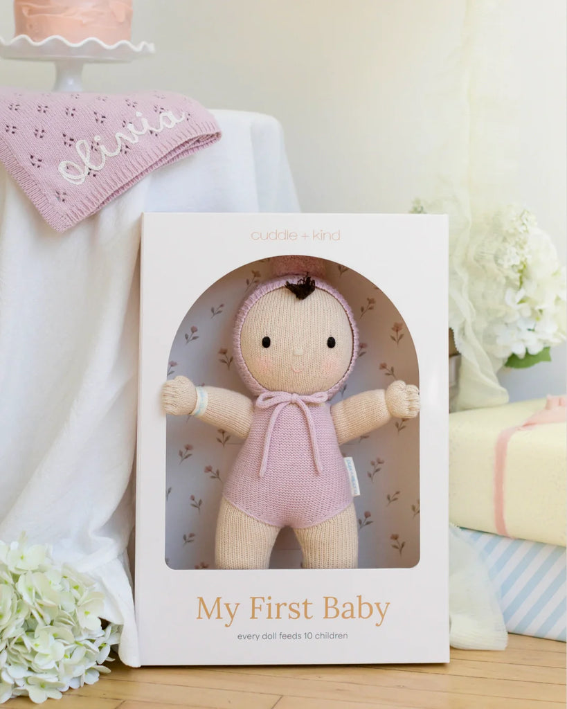 A boxed Cuddle + Kind My First Baby (Lavender) doll is displayed on a table. The doll wears a lavender outfit and bonnet. Near it are white flowers, a pink "Olivia" hat, and a pink cake on a stand. Ships in one week.