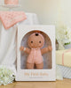 A soft, hand-knit Cuddle + Kind My First Baby (Peach) doll is displayed in a white box alongside white flowers, a pink cake, and a personalized pink blanket reading "emma". Ships in one week.