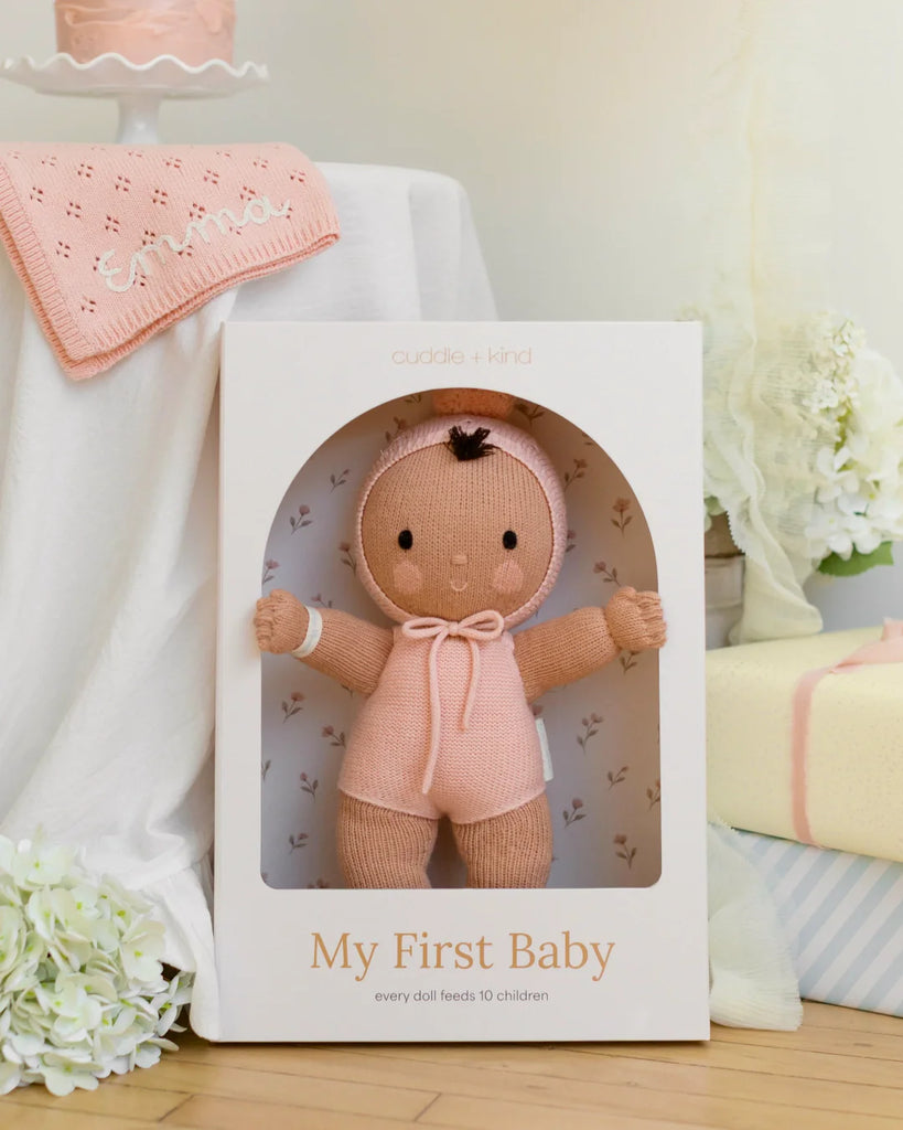 A soft, hand-knit Cuddle + Kind My First Baby (Peach) doll is displayed in a white box alongside white flowers, a pink cake, and a personalized pink blanket reading "emma". Ships in one week.