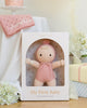 The Cuddle + Kind My First Baby (Rose) doll, hand-knit from natural cotton yarn in a pink outfit and hat, is displayed upright in pastel "My First Baby" packaging. Nearby are a pink personalized blanket, cake, flowers, and white fabric. Ships in one week.