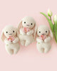Three heirloom-quality Cuddle + Kind Baby Bunny (Blooming Tulip) plush toys—cream-colored with pink ears—hold pink flowers on a light pink background beside two white tulips, all crafted from soft Peruvian cotton yarn.