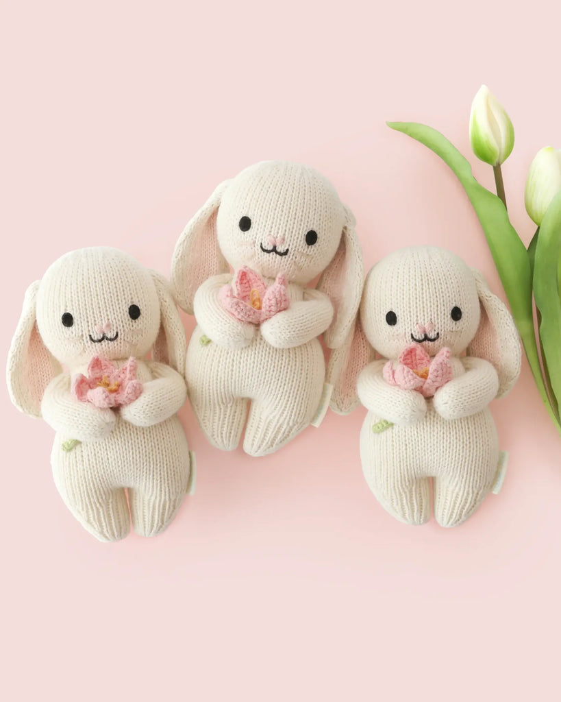 Three heirloom-quality Cuddle + Kind Baby Bunny (Blooming Tulip) plush toys—cream-colored with pink ears—hold pink flowers on a light pink background beside two white tulips, all crafted from soft Peruvian cotton yarn.