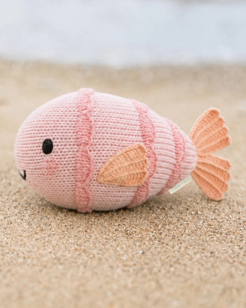 The Cuddle + Kind Baby Fish is a hand-knit pink plush toy with orange fins and tail, black eyes, and a small smile—perfect for cuddling and collecting.