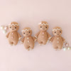 Four Cuddle + Kind Baby Gingerbread dolls, crafted from natural Peruvian cotton yarn, featuring smiling faces with white icing-like details and colorful buttons, are set against a soft pink background. Two pastel-colored ornaments complement these heirloom-quality pieces, positioned on either side.