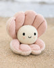 Cuddle + Kind Baby Pearl, a hand-knit heirloom plush shaped like a smiling pearl in a pink clam shell, sits on the sandy beach with blurred water behind.