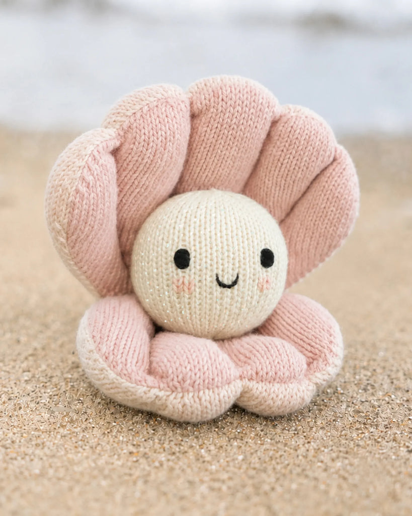 Cuddle + Kind Baby Pearl, a hand-knit heirloom plush shaped like a smiling pearl in a pink clam shell, sits on the sandy beach with blurred water behind.