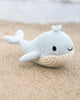 A Cuddle + Kind Baby Whale, hand-knit in Peru with a smiling face, rests on the sandy beach near the water.