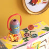 The Moulin Roty Storybook Lantern - The Minouchkas, featuring a red handle, projects images onto a yellow floral box. Three round image reels are shown—one in the lantern and two on the box—with framed art on the yellow wall.