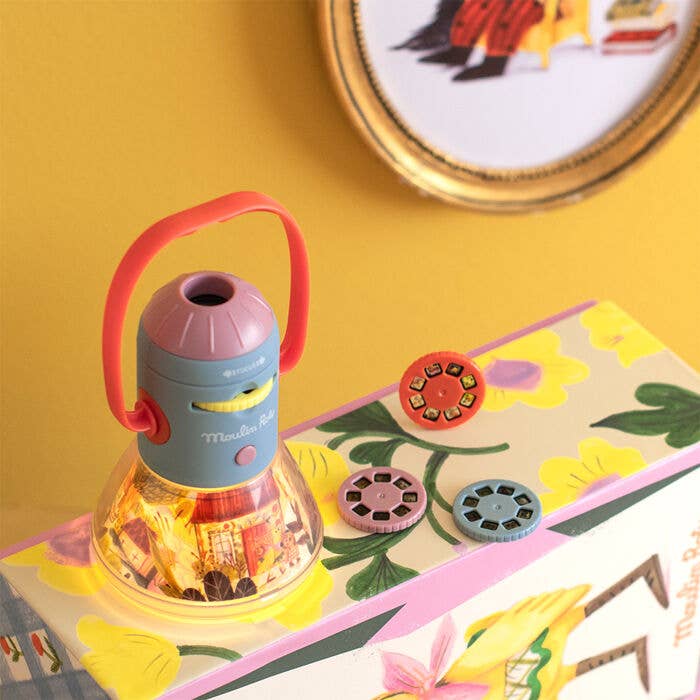 The Moulin Roty Storybook Lantern - The Minouchkas, featuring a red handle, projects images onto a yellow floral box. Three round image reels are shown—one in the lantern and two on the box—with framed art on the yellow wall.