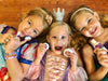 Three young girls in princess costumes lying on the floor, holding small containers of the Klee Naturals Deluxe Makeup Kit - Unicorn Cloud Fairy.