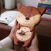 A person holds a Handmade Wooden Fox Wall Clock featuring a cute fox design with the clock face on its belly. Books and an open notebook rest on a wooden table—ideal whimsical decor for a nursery.