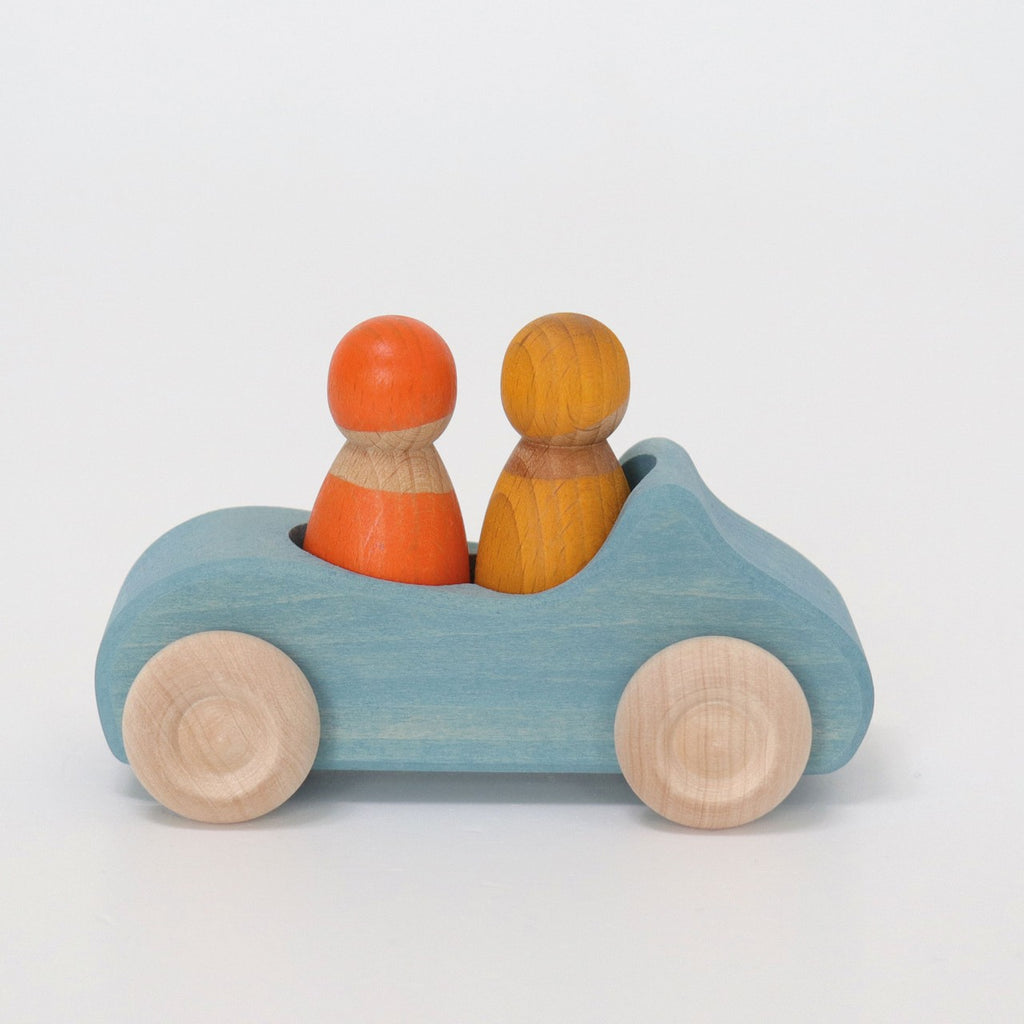 A Grimm's Wooden Blue Convertible Car painted light blue with non-toxic color stain features natural wood wheels. Two wooden peg figures are seated inside: one painted orange and the other yellow. The car and figures, part of the Rainbow Friends toy line, are against a plain white background.