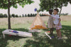 Two children play a Cornhole Game in a sunny outdoor setting with grass and trees. A cream-colored tent with yellow interior fabric is set up behind them, and wooden blocks are scattered on the ground. The Cornhole Game features all-weather bean bags and a regulation-sized cornhole board, set against a backdrop of a meadow and distant trees.