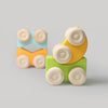 Two vibrant Grimm's Stacking Cars are placed on a light gray surface—one is the green (coming soon) model with yellow accents, while the other is blue and orange with a distinctive square shape in the background.