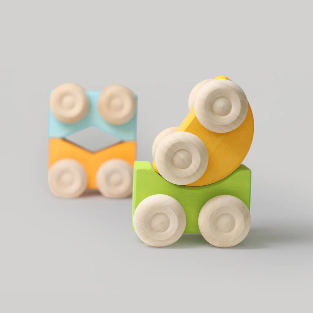 Two vibrant Grimm's Stacking Cars are placed on a light gray surface—one is the green (coming soon) model with yellow accents, while the other is blue and orange with a distinctive square shape in the background.