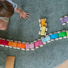 A child sits on a green carpet, playing with Grimm's Stacking Cars - Pink arranged in a line. The child's arm and legs are visible from above.