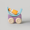 Grimm's Stacking Cars - Purple (coming soon): A minimalist wooden stacking toy with a purple rectangular base on two wheels, a blue semicircular cradle, and a yellow and white figure inside, set against a plain gray background.
