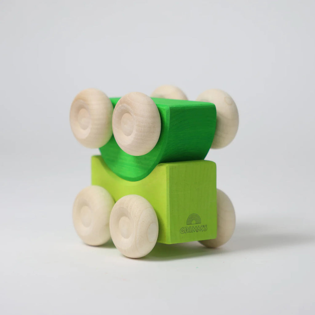 Grimm's Stacking Cars - Green (coming soon) features a green wooden car with six natural wood wheels in two stackable layers, set on a plain white background. The bottom block displays the engraved "Grimm's" logo.