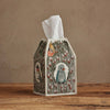 The Coral & Tusk Birdhouse Tissue Box Cover, featuring embroidered birds, butterflies, and florals, rests on a wooden surface against a beige wall with a white tissue peeking from the top.