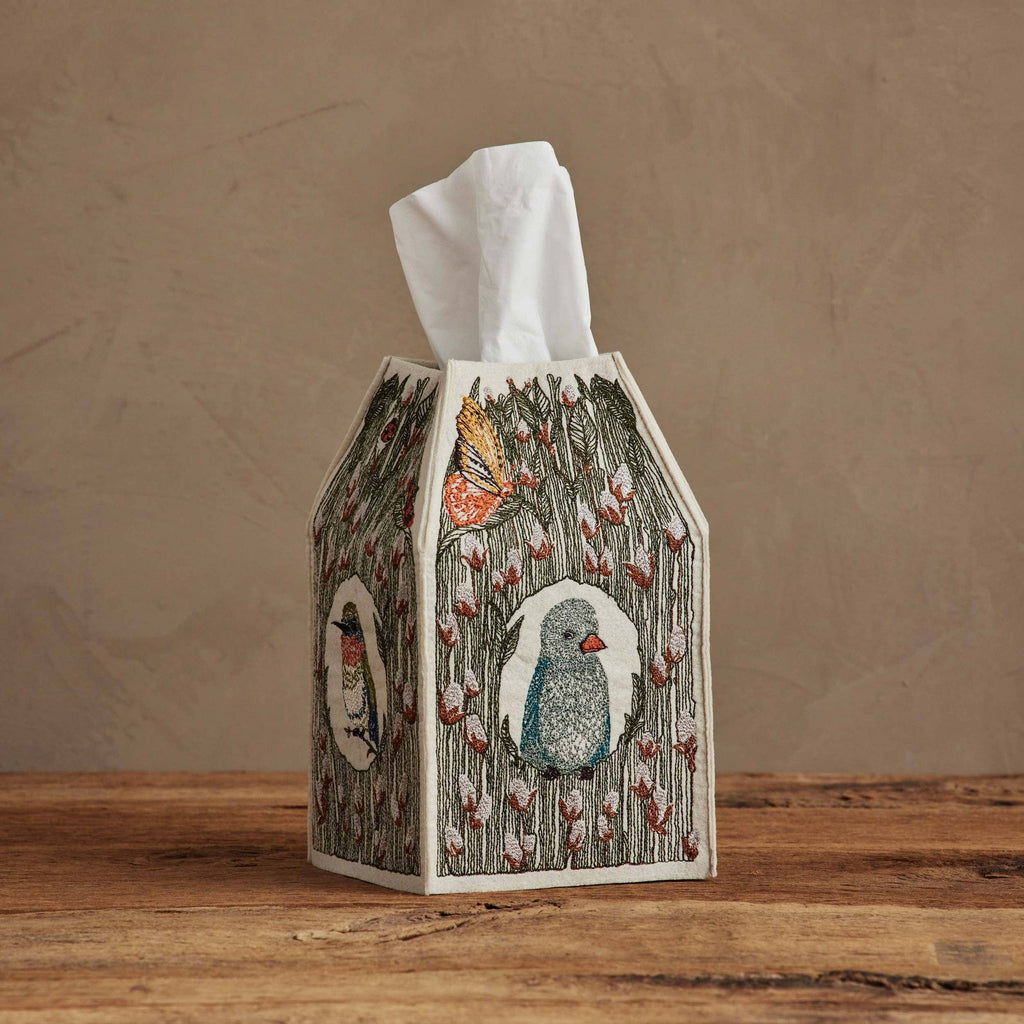 The Coral & Tusk Birdhouse Tissue Box Cover, featuring embroidered birds, butterflies, and florals, rests on a wooden surface against a beige wall with a white tissue peeking from the top.